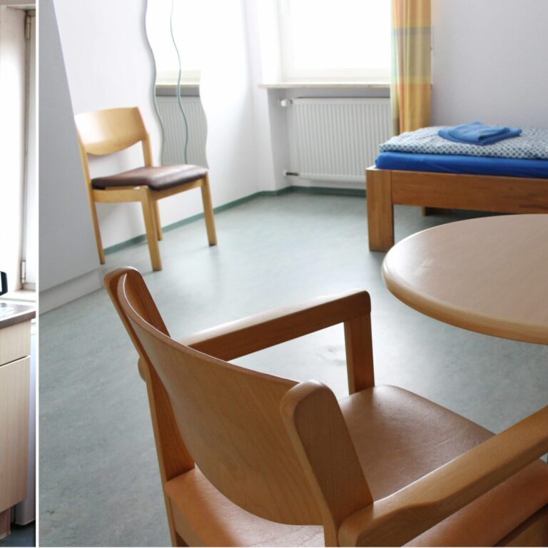 Diashow Bild eines Stationären Zimmers. Auf der linken Seite ist eine helle Küche zu sehen mit Spülbecken und Kaffeemaschine und auf der rechten Seite ist ein Schlafzimmer mit Bett, Spiegel, einem kleinen runden Tisch und Stuhl zu sehen.