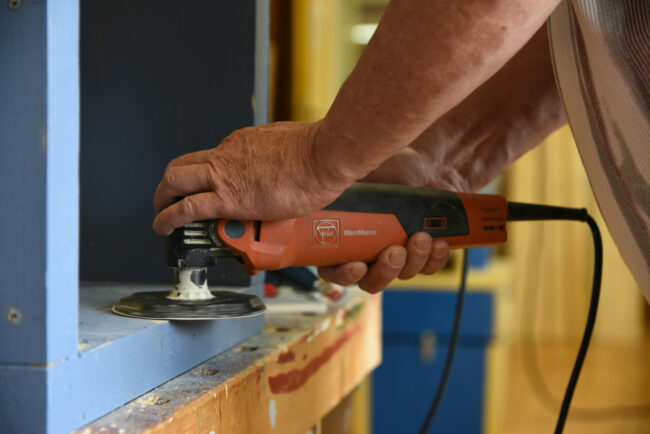Nahaufnahme einer Person beim Schleifen eines Holzstücks mit einem elektrischen Schleifgerät, in einer Werkstattumgebung.