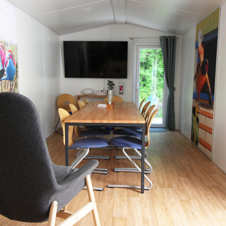 In einem Gemeinschaftszimmer steht ein großer Tisch mit mehreren Stühlen. Im Hintergrund hängt ein Fernseher an der Wand und an den Wänden links und rechts hängen 2 große Bilder. Vorne steht ein grauer Sesselstuhl, der zum Fernseher gedreht ist.