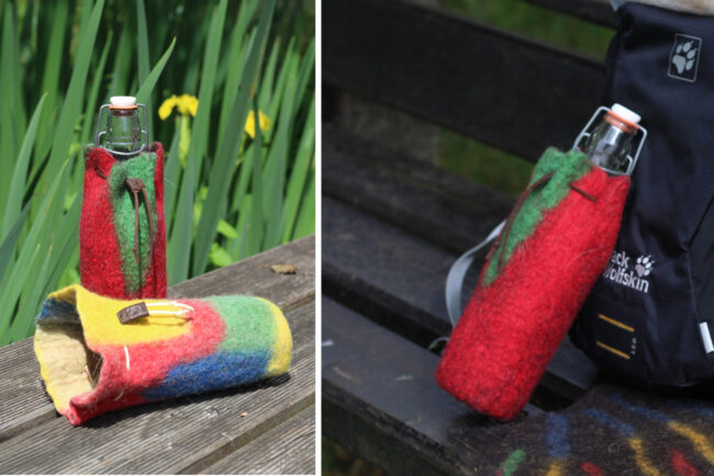 Zwei bunte Filzhüllen für Trinkflaschen – eine aufrecht mit Flasche, eine liegend – liegen auf einer Holzbank am Wasser. Rechts im Bild ist dieselbe Hülle an einem Rucksack befestigt.
