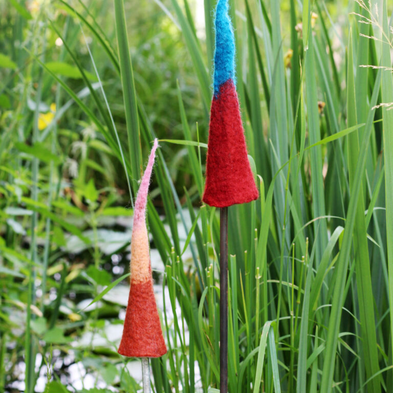 Zwei bunte Filzobjekte in Form spitzer Hüte stecken an Metallstangen im hohen Gras neben einem Teich – kunstvolle Gartendekoration.