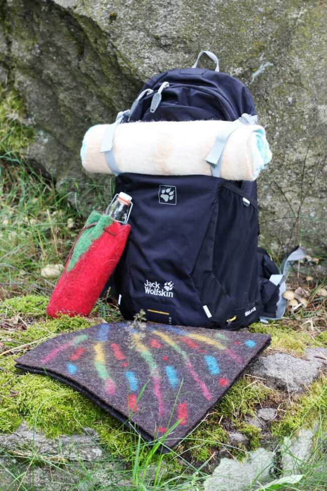 in dunkler Wanderrucksack mit aufgerollter Decke, einer roten Filzhülle für eine Trinkflasche und einer bunten Sitzmatte liegt vor einem großen Felsen im Moos.
