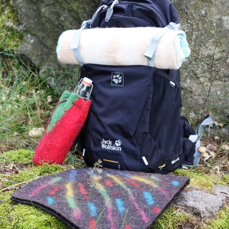 in dunkler Wanderrucksack mit aufgerollter Decke, einer roten Filzhülle für eine Trinkflasche und einer bunten Sitzmatte liegt vor einem großen Felsen im Moos.