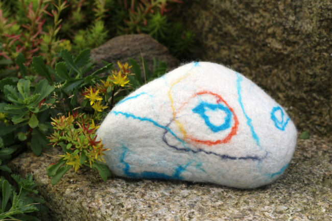Ein weiß umfilzter Stein mit abstrakten, bunten Linien liegt dekorativ zwischen blühenden Sukkulenten und Felsen im Garten.