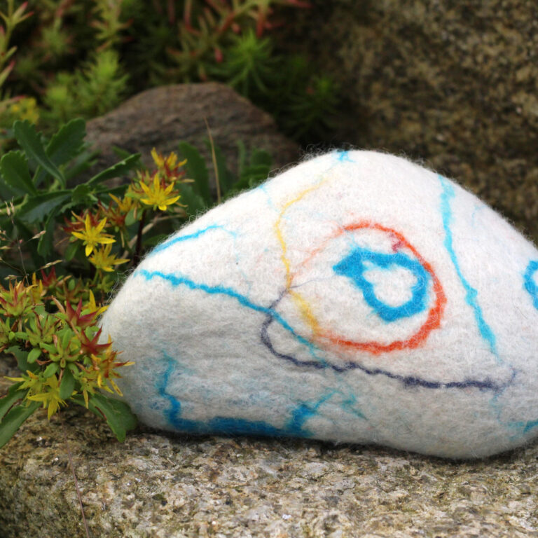Ein weiß umfilzter Stein mit abstrakten, bunten Linien liegt dekorativ zwischen blühenden Sukkulenten und Felsen im Garten.