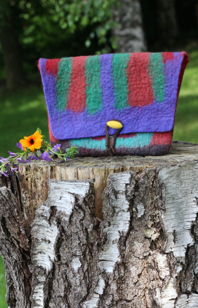 Eine bunt gestreifte Filztasche in kräftigem Rot, Lila, Grün und Türkis steht auf einem Baumstumpf. Daneben eine kleine orangefarbene Blume mit violetten Blüten.