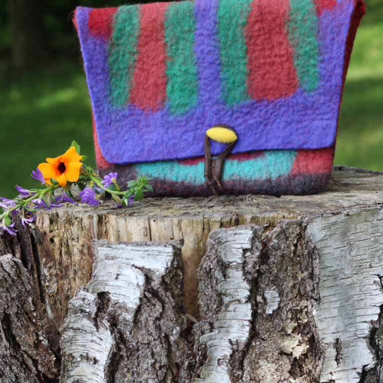 Eine bunt gestreifte Filztasche in kräftigem Rot, Lila, Grün und Türkis steht auf einem Baumstumpf. Daneben eine kleine orangefarbene Blume mit violetten Blüten.