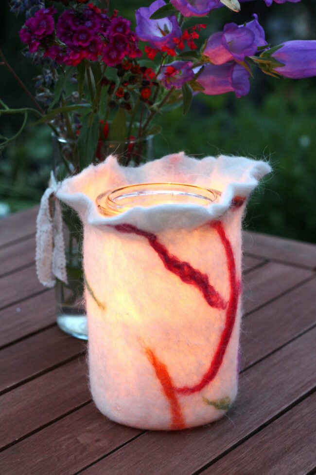 Ein Windlicht aus weißem, leicht durchscheinendem Filz mit roten und orangen Linienmustern leuchtet warm auf einem Holztisch. Dahinter ein Glas mit buntem Wiesenblumenstrauß.