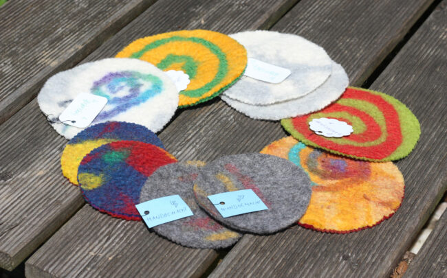 Mehrere bunte, runde Filzuntersetzer mit Spiralen und Mustern liegen kreisförmig auf einer Holzbank. Kleine Etiketten mit der Aufschrift „Handgemacht“ sind an einigen befestigt.