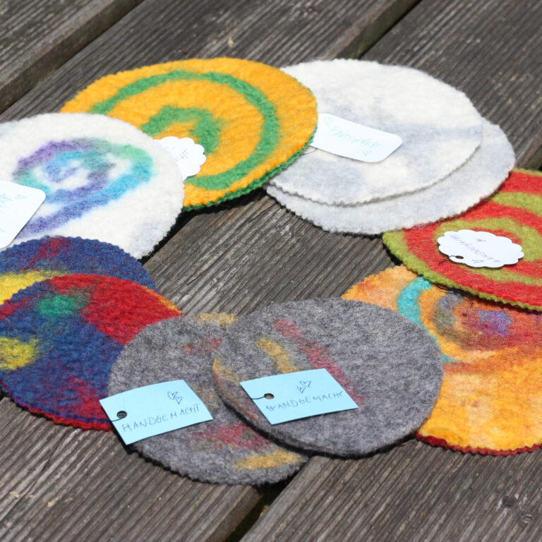 Mehrere bunte, runde Filzuntersetzer mit Spiralen und Mustern liegen kreisförmig auf einer Holzbank. Kleine Etiketten mit der Aufschrift „Handgemacht“ sind an einigen befestigt.
