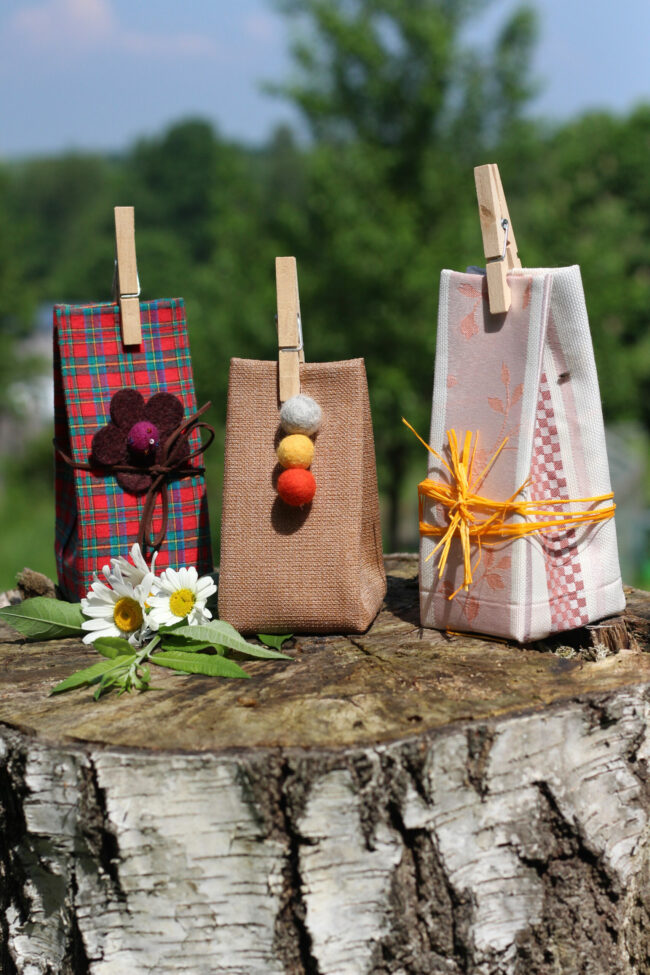 Drei Geschenkverpackungen aus Stoff mit liebevoller Dekoration – Filzblumen, Filzkugeln und Bastschleife – stehen auf einem Baumstumpf. Davor liegen zwei Gänseblümchen.