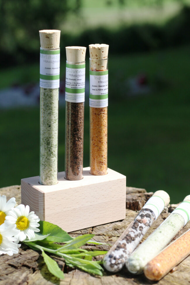 Drei Glasröhrchen mit verschiedenfarbigen Gewürzmischungen stehen in einem Holzblock. Weitere Röhrchen und Gänseblümchen liegen daneben auf dem Baumstumpf.