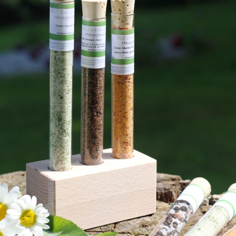 Drei Glasröhrchen mit verschiedenfarbigen Gewürzmischungen stehen in einem Holzblock. Weitere Röhrchen und Gänseblümchen liegen daneben auf dem Baumstumpf.