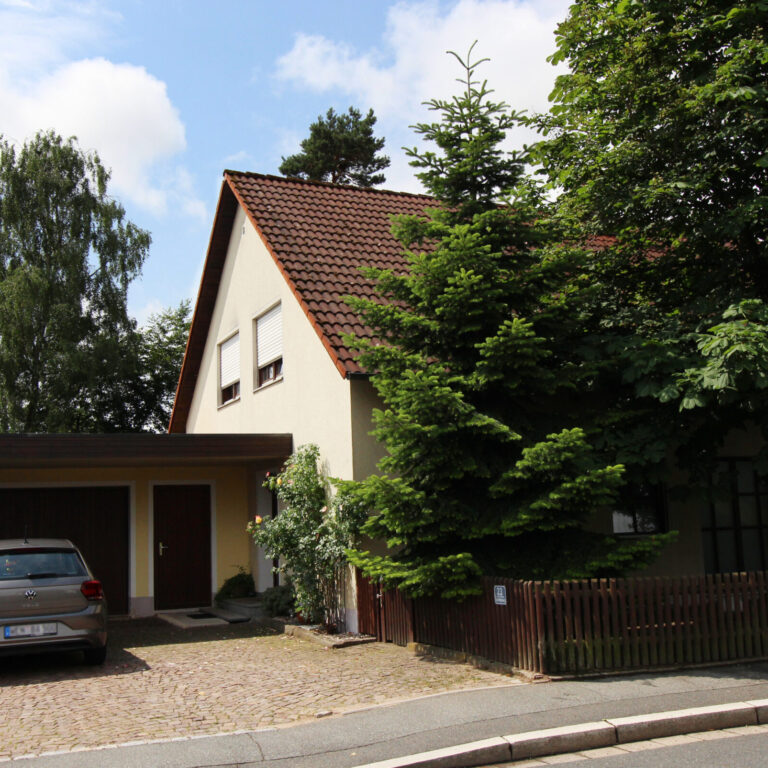 Zweistöckiges Wohnhaus mit Satteldach, Carport, geparktem Auto und Vorgarten mit Bäumen – Ansicht von der Straße