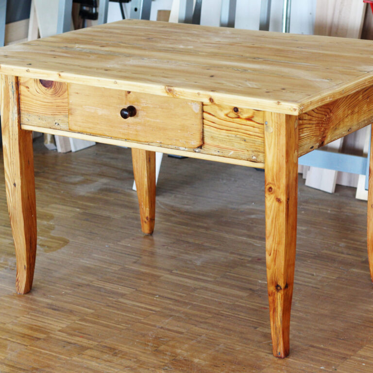 Ein massiver Holztisch mit Schublade, abgeschliffener Oberfläche und rustikalem Charme steht in einer Werkstatt mit sichtbaren Arbeitsmaterialien im Hintergrund.