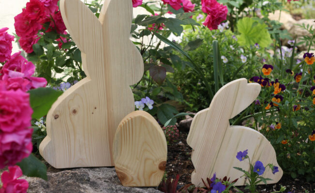 Drei Hasenfiguren aus hellem Holz stehen zwischen bunten Blumen in einem Beet.