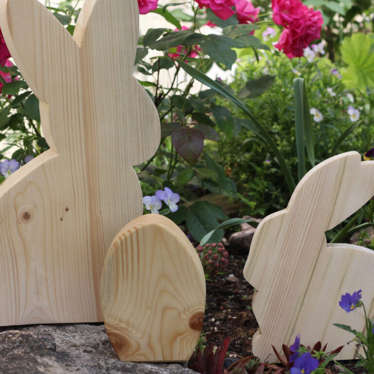 Drei Hasenfiguren aus hellem Holz stehen zwischen bunten Blumen in einem Beet.