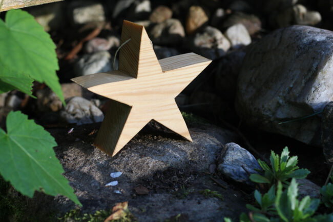 Ein dekorativer Holzstern mit Aufhängung steht auf einem Stein im Garten, umgeben von natürlichen Steinen und Blattgrün.