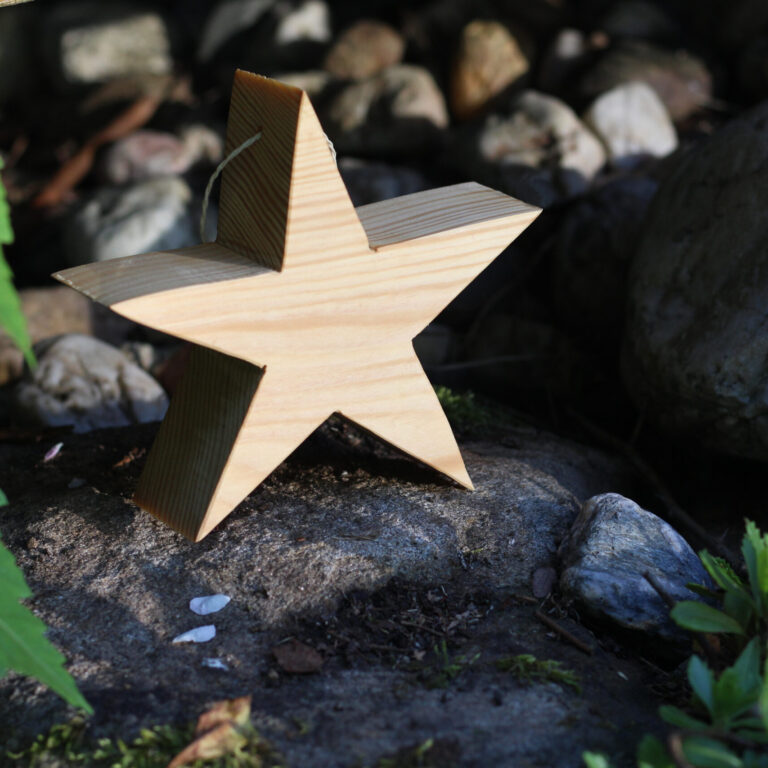 Ein dekorativer Holzstern mit Aufhängung steht auf einem Stein im Garten, umgeben von natürlichen Steinen und Blattgrün.