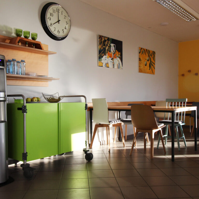 Ein Speisesaal, in dem mehrere Tische und verschiedene Stühle stehen. Links ist ein Wasserautomat und ein Grüner Schrank.