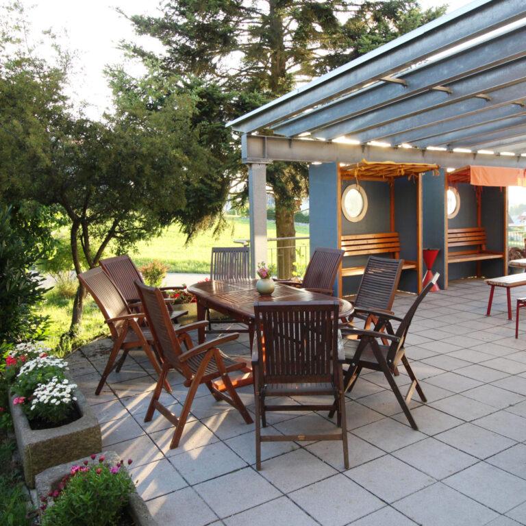 Eine große Terrasse, auf der mehrere Tische und Stühle stehen. Sie ist überdacht und es gibt viele Sitzmöglichkeiten.
