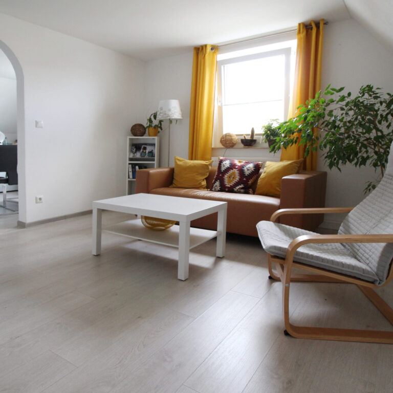 Gemütliches Wohnzimmer mit braunem Sofa, weißem Couchtisch, Sessel und gelben Vorhängen – im Hintergrund Zugang zur Küche mit Esstisch.