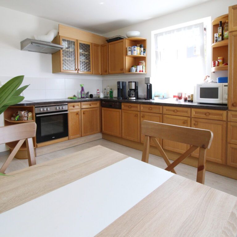 Moderne Küche mit Holzfronten, Esstisch, Elektrogeräten und großem Fenster – gemütlicher Raum für gemeinsames Kochen und Essen.