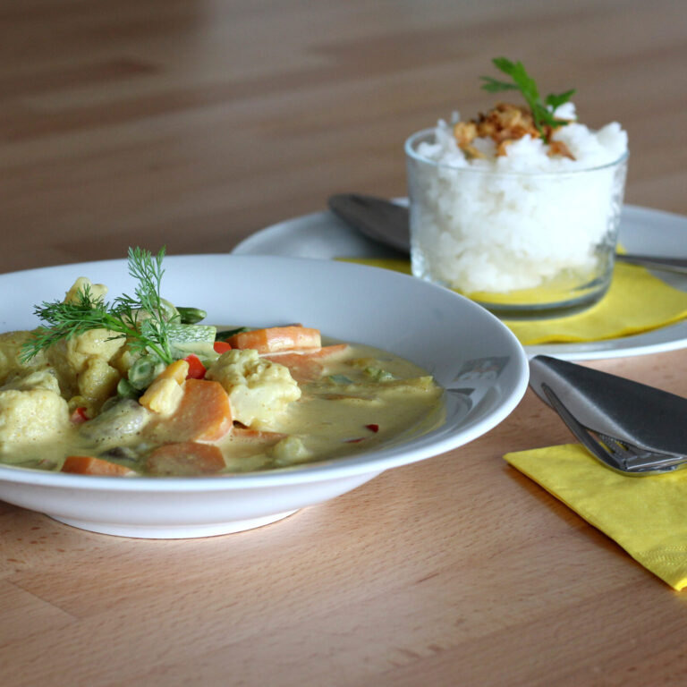 Gesundes Mittagessen in der Klinik: Teller mit Gemüse-Curry und Glas mit Reis, serviert auf hellem Holztisch mit Besteck und gelber Serviette.