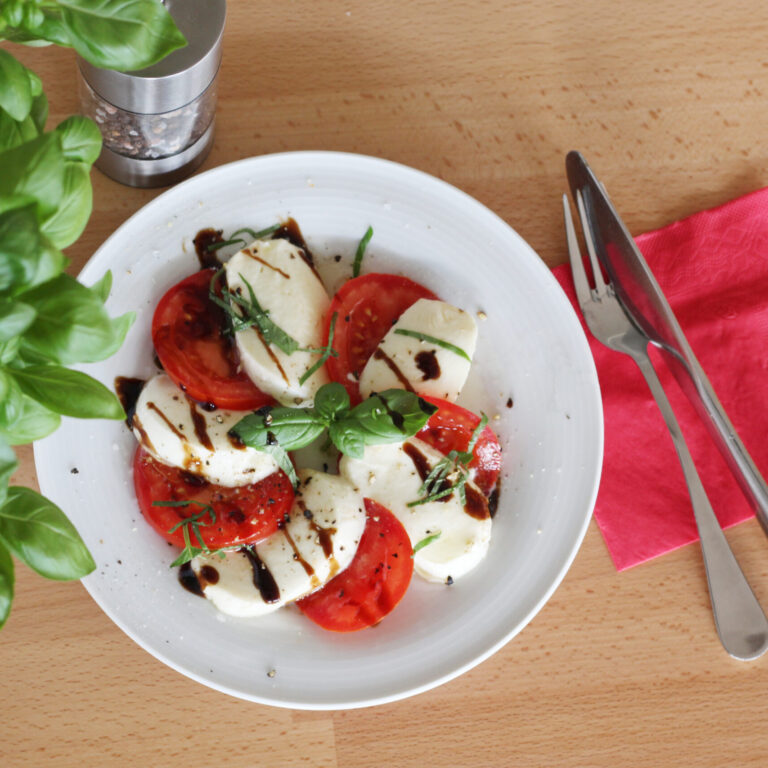 Tomate Mozzarella, serviert mit Basilikum auf hellem Holztisch mit Besteck und roter Serviette.