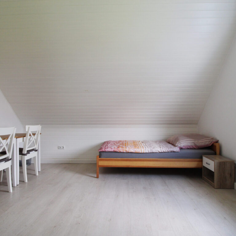 Minimalistisch eingerichtetes Zimmer mit Einzelbett, weißem Kleiderschrank, Fenster, Schreibtisch und zwei weißen Stühlen.