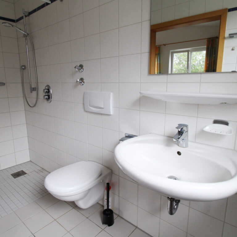 Stationäres Badezimmer. Helle Fliesen an der Wand, offene Dusche, daneben eine Toilette, ein Waschbecken und darüber ein Spiegel.