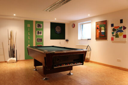 Ein heller Freizeitraum mit Billardtisch, Dartboard und mehreren gerahmten Bildern an der Wand. Daneben ein Halter mit Billard-Queues.