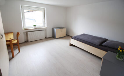 WG Zimmer in Weiden, das ein Dunkles Bett, einen farblich passenden Nachttisch, Schrank und einen Tisch enthält.