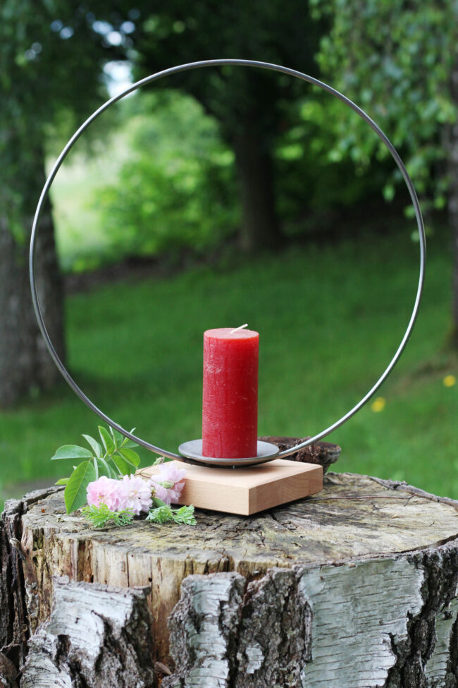 Eine rote Stumpenkerze steht in einem modernen Kerzenhalter mit rundem Metallrahmen auf einem Baumstumpf, verziert mit rosa Blüten.