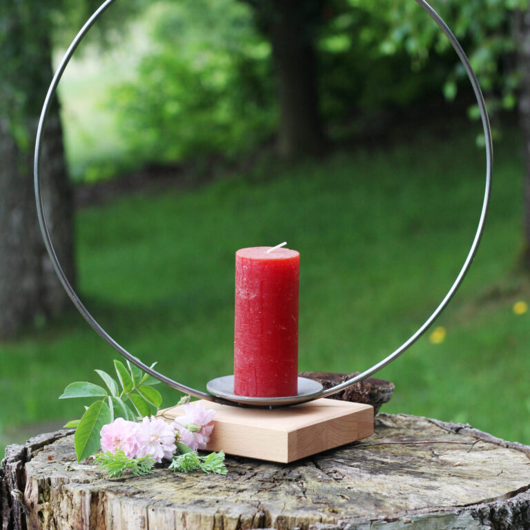 Eine rote Stumpenkerze steht in einem modernen Kerzenhalter mit rundem Metallrahmen auf einem Baumstumpf, verziert mit rosa Blüten.
