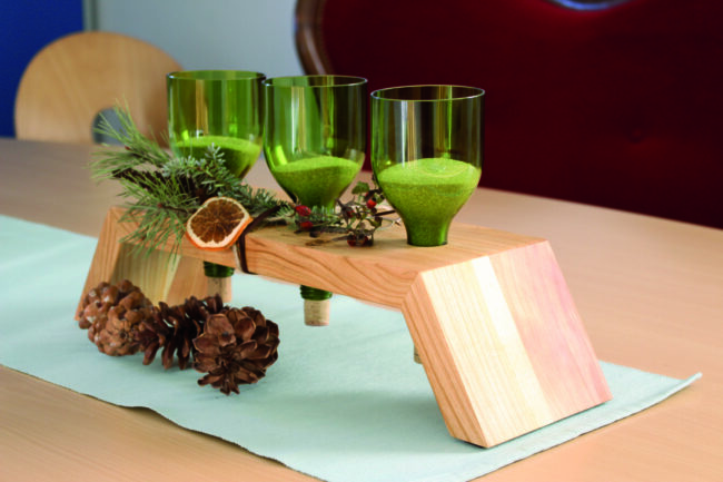 Weihnachtliche Deko auf Holztisch. Die Deko zeigt ein Holzpodest, in das abgeschnittene Grüne Flaschen gesteckt wurden. Diese sind mit Sand gefüllt und Rundherum sind Tannenzweige und Orangenscheiben