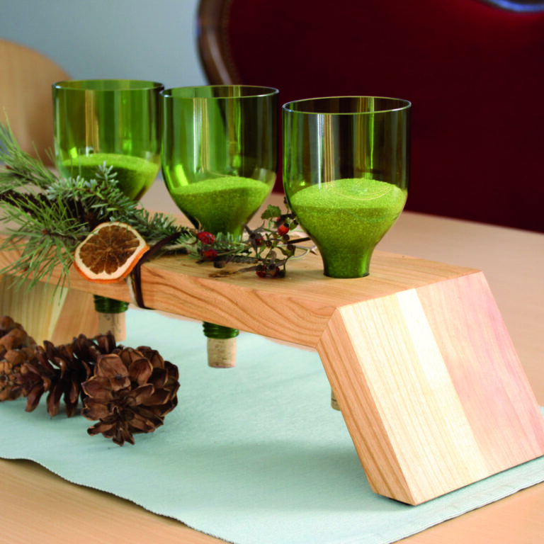 Weihnachtliche Deko auf Holztisch. Die Deko zeigt ein Holzpodest, in das abgeschnittene Grüne Flaschen gesteckt wurden. Diese sind mit Sand gefüllt und Rundherum sind Tannenzweige und Orangenscheiben