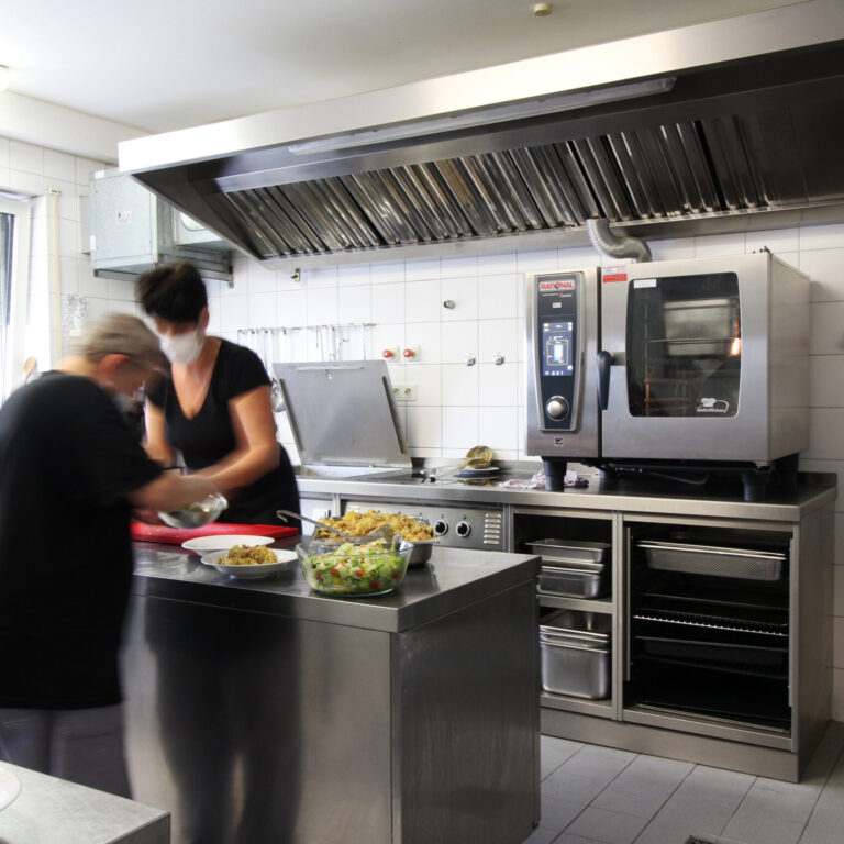 Zwei Frauen tragen einen Mundschutz und stehen in einer Küche um zu Kochen. Um sie herum stehen mehrere Schüsseln mit Salat und Kartoffeln.
