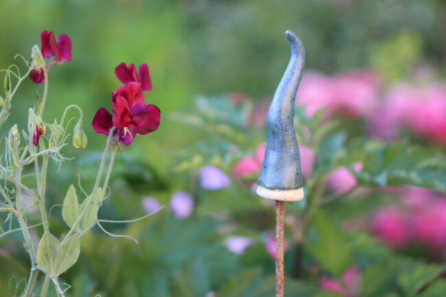 Zipfelmütze aus Keramik sitzt auf einem Metallstab, in einem grünen Garten, neben Roten Blüten.