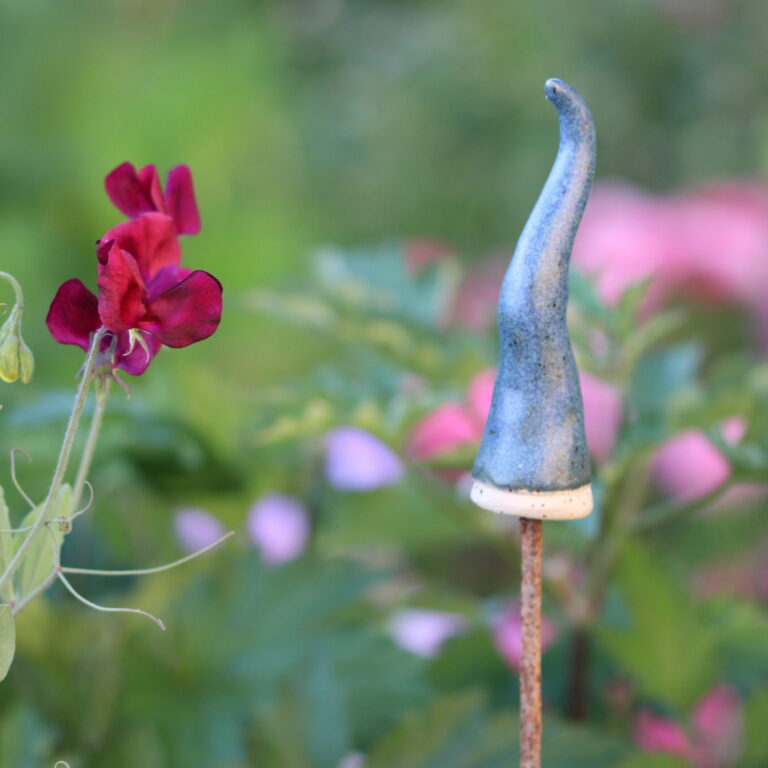 Zipfelmütze aus Keramik sitzt auf einem Metallstab, in einem grünen Garten, neben Roten Blüten.