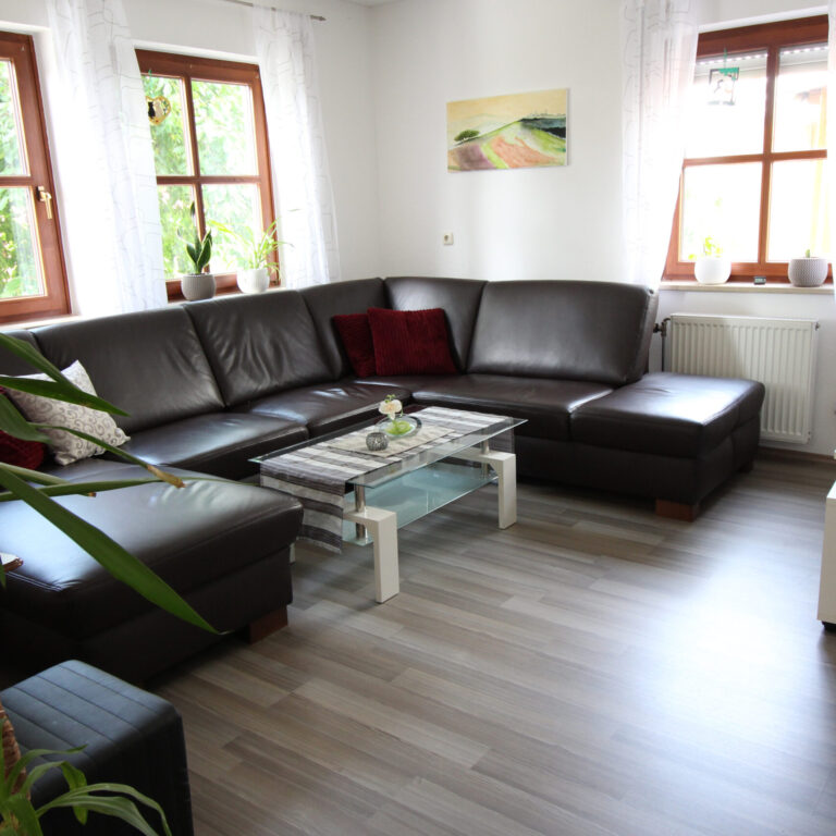 Großes Wohnzimmer mit dunkelbraunem Ecksofa, Couchtisch, Fernseher und drei Fenstern mit weißen Vorhängen – helle und gemütliche Atmosphäre.