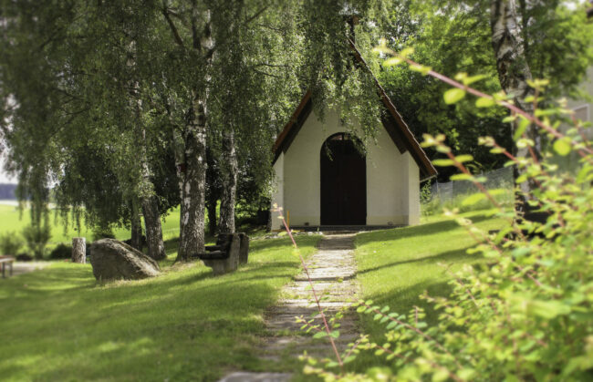 Ein Steinweg führt zu einer kleinen Kapelle, die in einem Sonnigen Garten steht. Neben der Kapelle stehen einige Bäume und eine Holzbank.