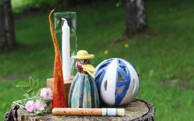 Ein Ensemble aus Kerze, Filzfigur, Keramikhuhn mit Hut, Keramikkugel, Blumen und Glasröhrchen auf einem alten Baumstumpf im Grünen.