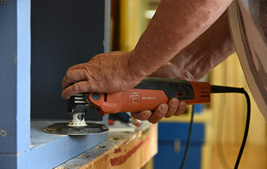 Nahaufnahme einer Person beim Schleifen eines Holzstücks mit einem elektrischen Schleifgerät, in einer Werkstattumgebung.