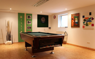 Ein heller Freizeitraum mit Billardtisch, Dartboard und mehreren gerahmten Bildern an der Wand. Daneben ein Halter mit Billard-Queues.